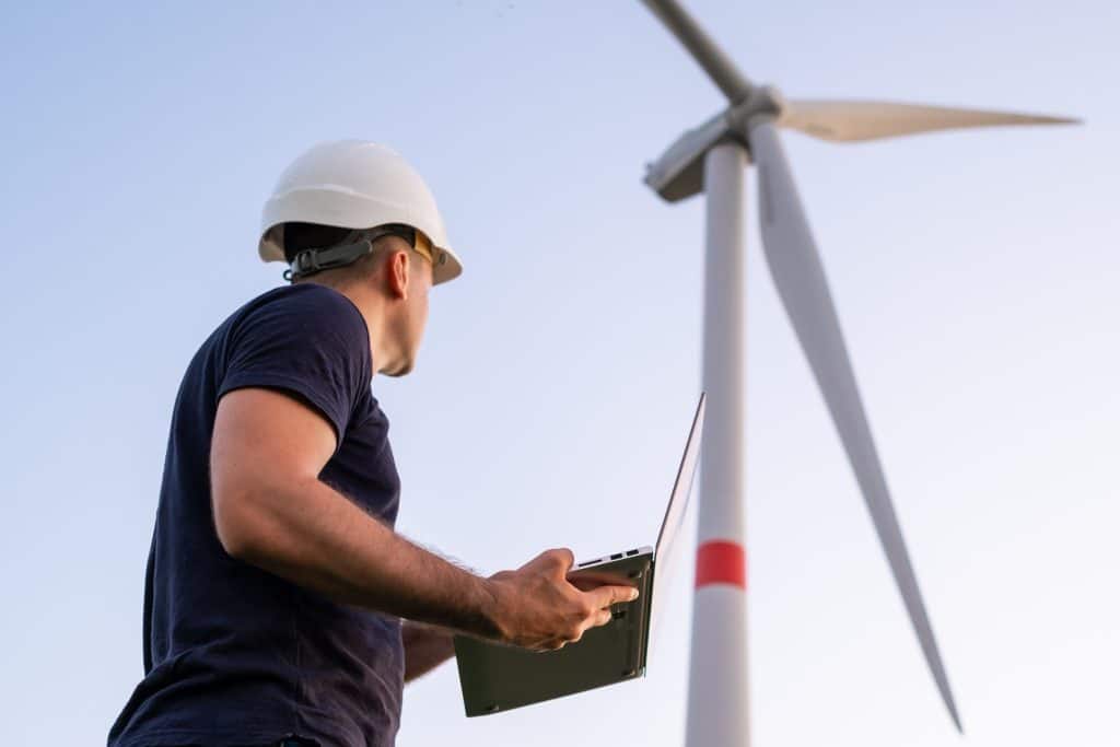 Male engineer in the helmet controls and maintains the operation of a wind turbine using a laptop on the background of windmill