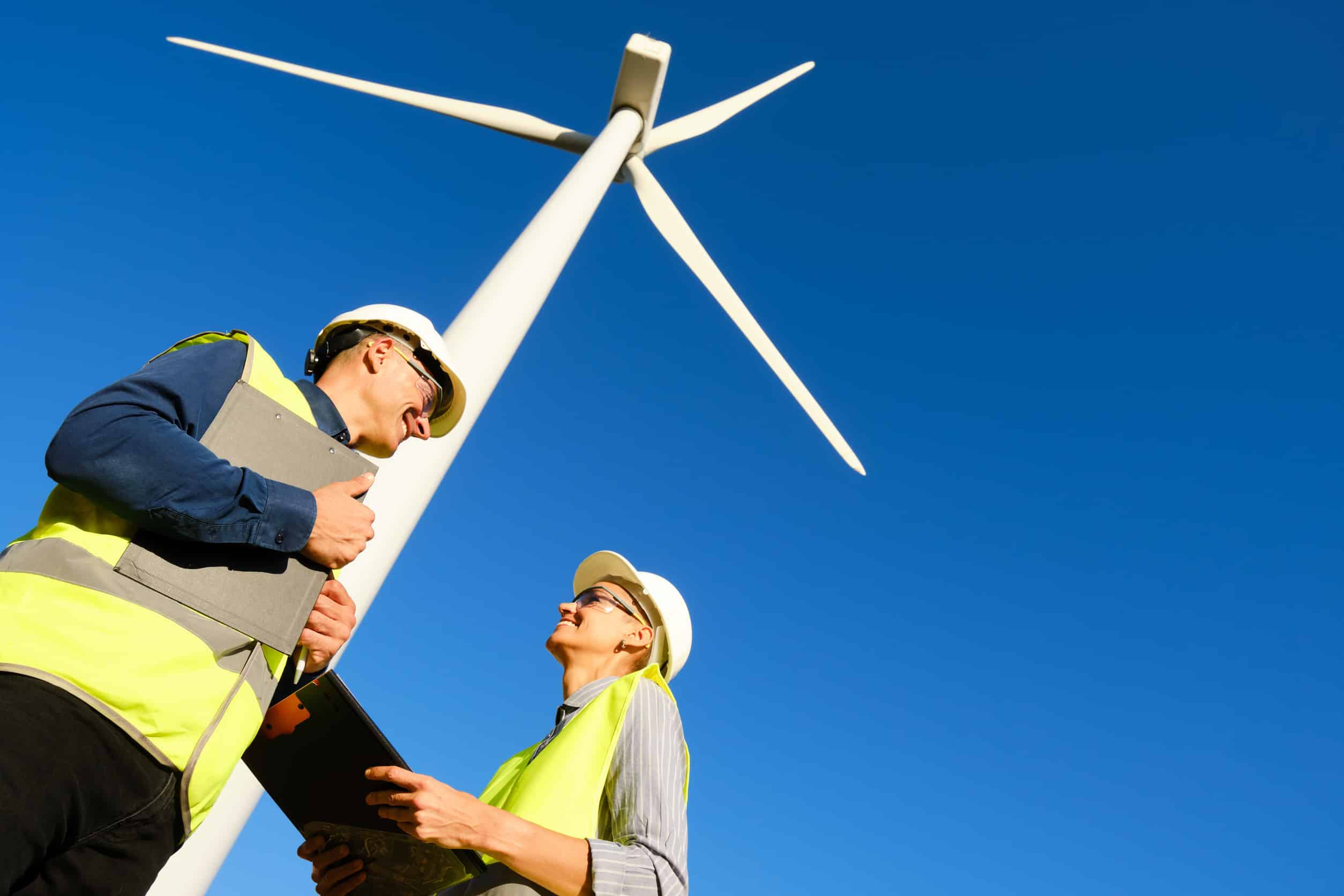 Two engineers wearing safety helmets and high-visibility vests inspecting a wind turbine at a renewable energy site under a clear blue sky.