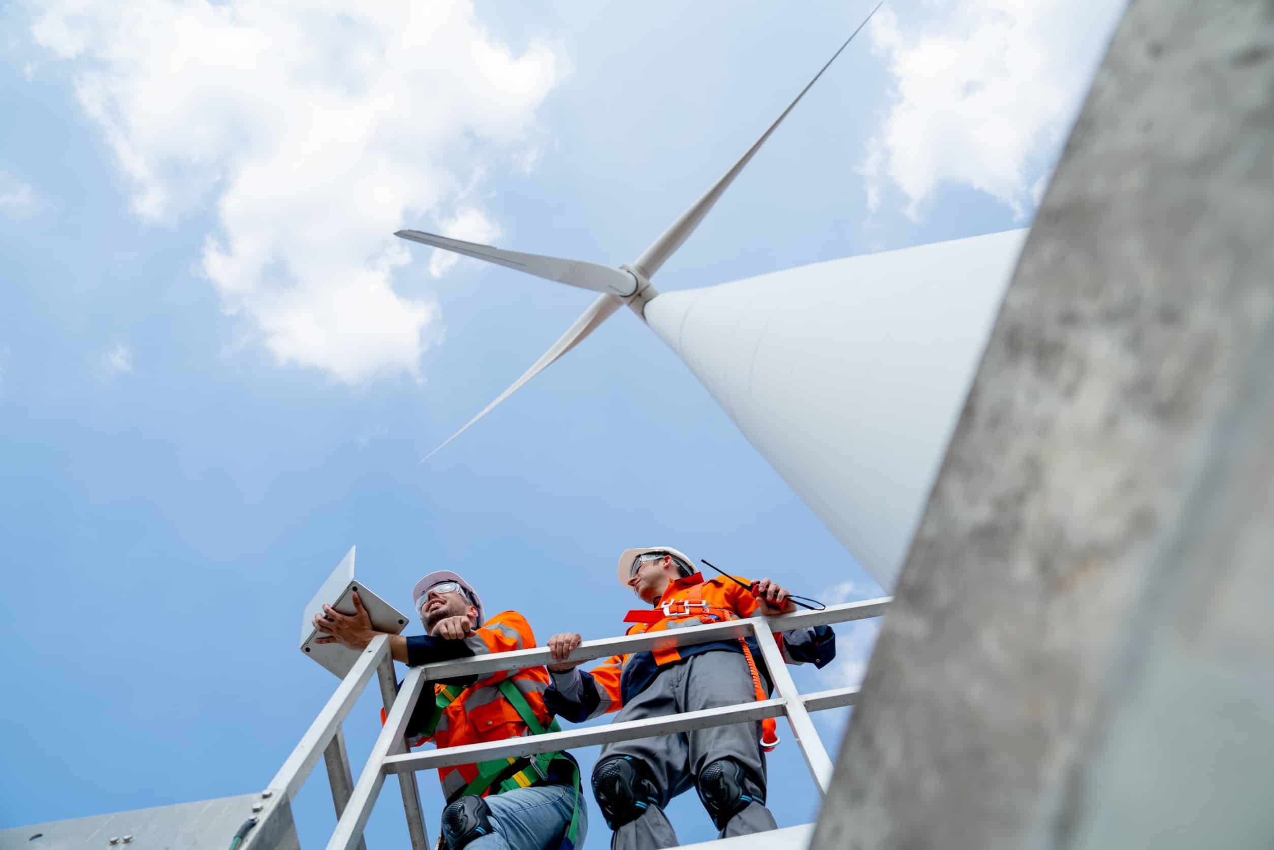 Two technicians in safety gear working on a wind turbine platform, using a tablet and radio for inspection and maintenance.