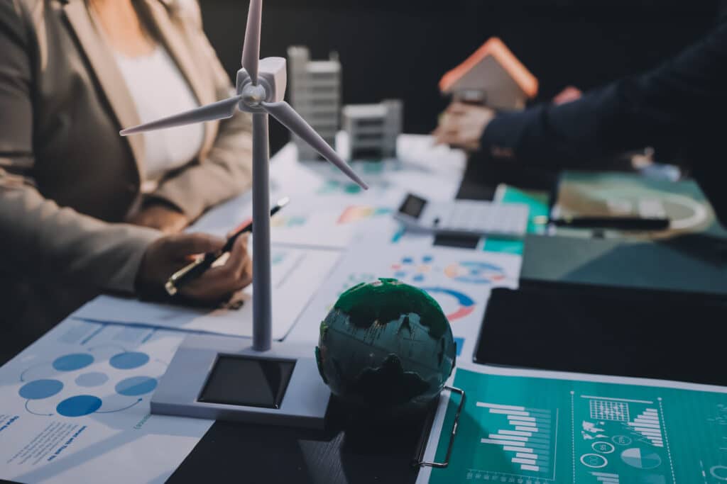 Close-up of a wind turbine model and globe on a desk with charts and documents, as professionals discuss sustainable energy and eco-friendly planning in the background.