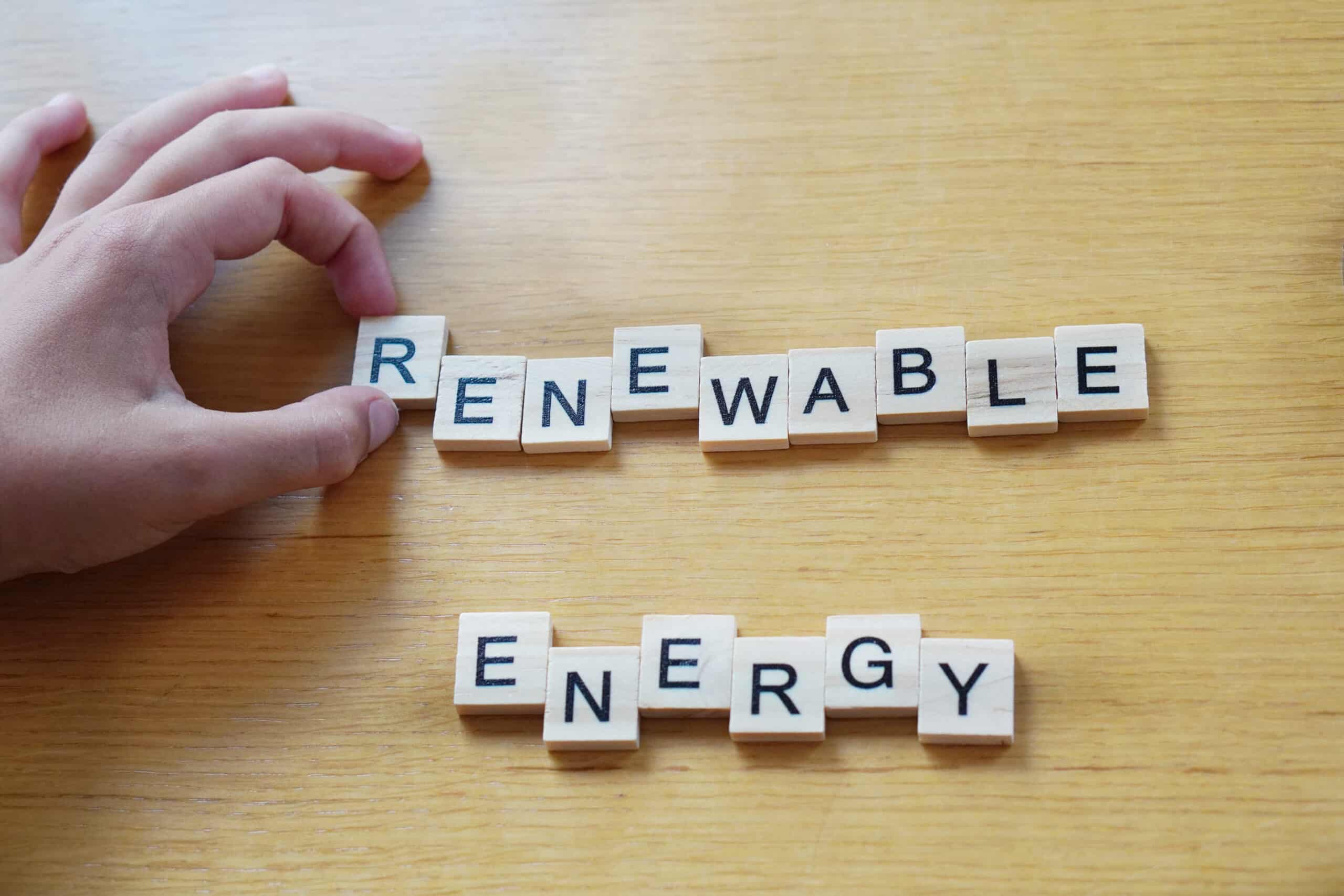 Hand arranging wooden letter tiles spelling “RENEWABLE ENERGY” on a wooden surface.
