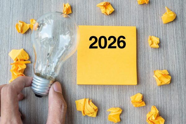 Hand holding a light bulb next to a yellow sticky note labeled “2026,” surrounded by crumpled paper balls on a wooden surface, symbolizing ideas and planning.